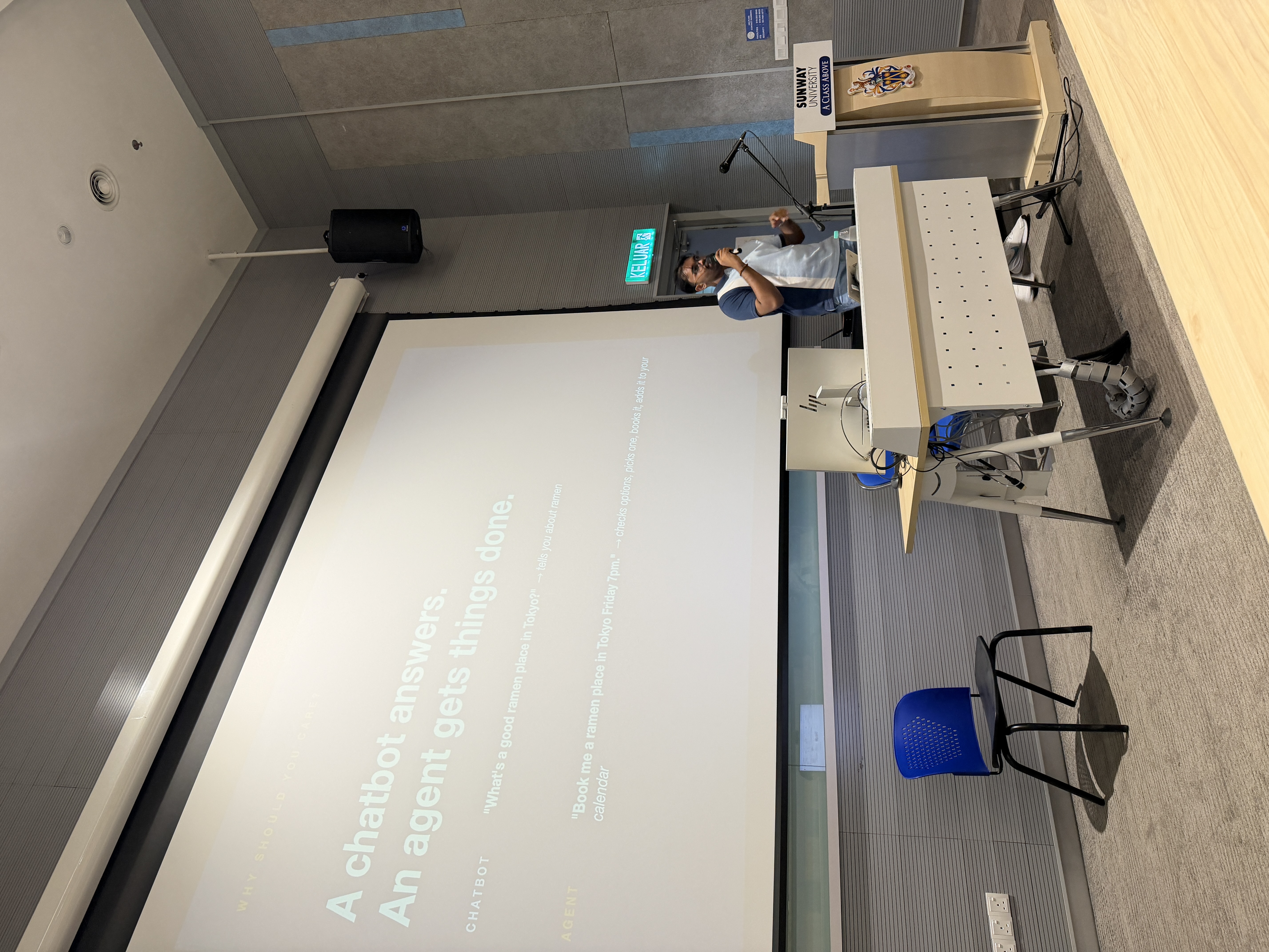 Slide reading 'A chatbot answers. An agent gets things done.' on the lecture theatre screen